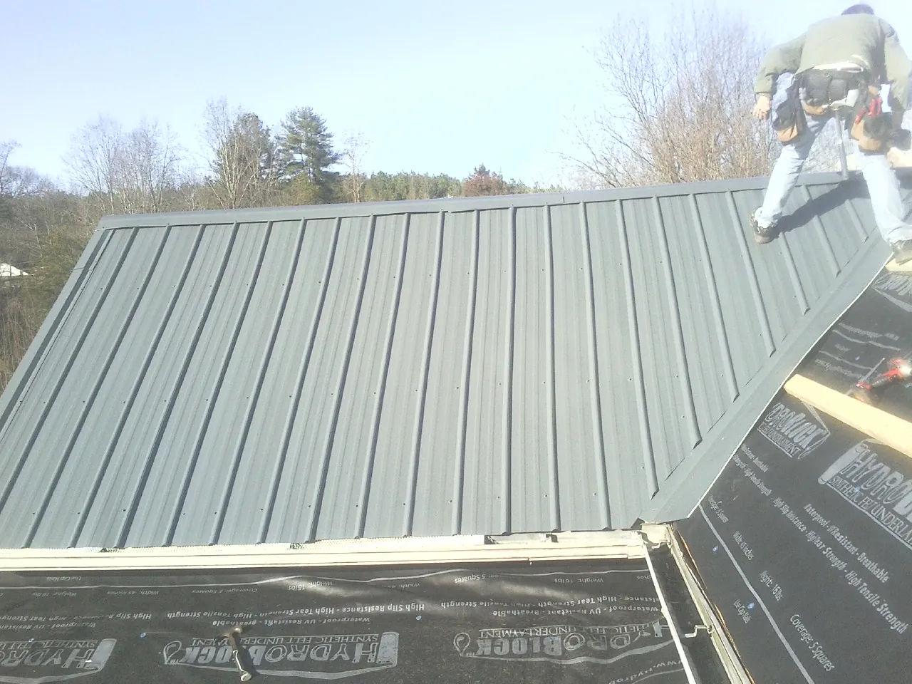 Crew installing dark green standing seam panels over synthetic underlayment in Marianna